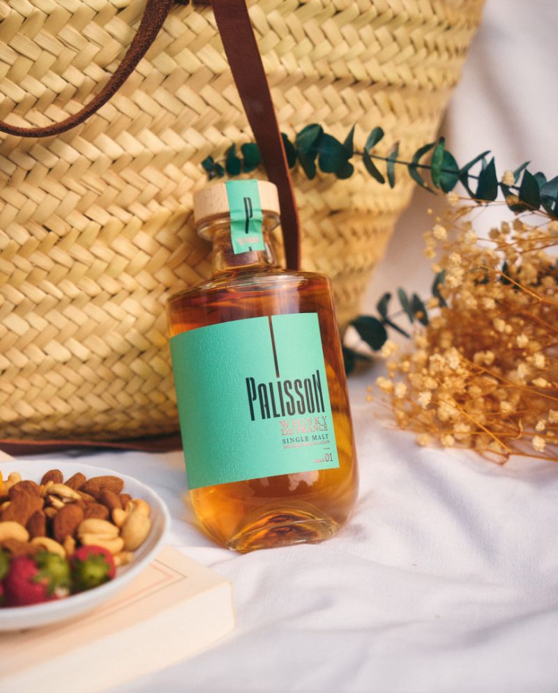 Photo de Palisson batch 01 whisky de France single malt finition en fûts de Cognac, devant un sac à main en osier avec des fleurs séchées, des noix et des fraises